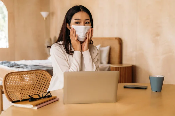 Jovem adolescente mulher asiática usa suéter usando laptop computador trabalhando em casa dentro de casa online. Menina atraente removendo uma máscara facial. — Fotografia de Stock