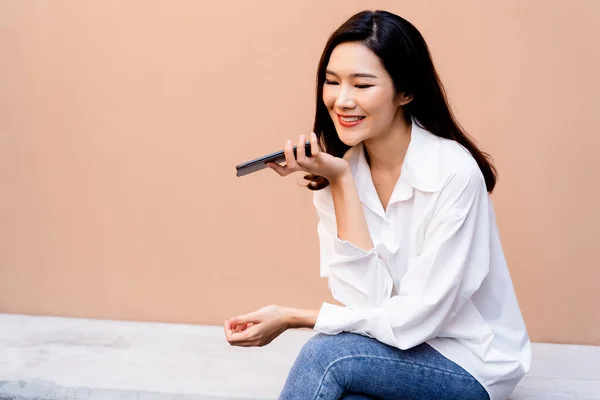 白いシャツとジーンズを着て背景に砂漠の砂の色の壁とコンクリートのベンチに座っている間、スマートフォンで会話をしている若い笑顔アジアの女の子 — ストック写真