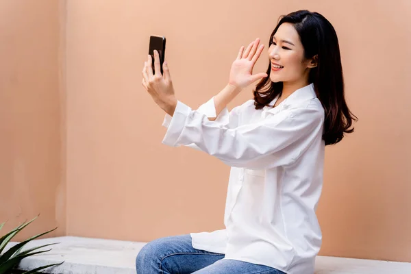 Gadis Asia muda dengan lipstik merah terang mengambil selfie dengan smartphone memiliki gurun pasir berwarna dinding di latar belakangnya mengenakan kemeja putih dengan tombol terbuka dan gaya rambut terbuka — Stok Foto