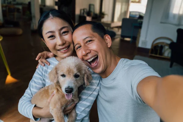 Joven pareja asiática adulta sosteniendo un cachorro tomando una selfie de un teléfono con el interior del hogar en el fondo. 30s maduro hombre y mujer con perro mascota tomando una foto de la familia. - Retrato de grupo feliz. —  Fotos de Stock