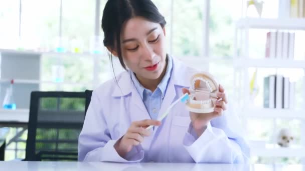 Portrét krásné mladé asijské zubařky demonstrující správný způsob, jak kartáč zuby drží ústa model a zubní kartáček při nahrávání video blog pro on-line výuku — Stock video