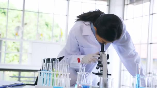 Jovem cientista asiática vestindo labcoat médico realizando pesquisa em laboratório enquanto vê elementos através de microscópio com equipamentos como tubo de ensaio e frasco — Vídeo de Stock