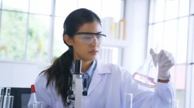Eczane laboratuarında çalışan genç bayan Asyalı tıp araştırmacısı tüpteki kimyasalları mikroskop ve test tüpü gibi aletlerle karıştırıyor.