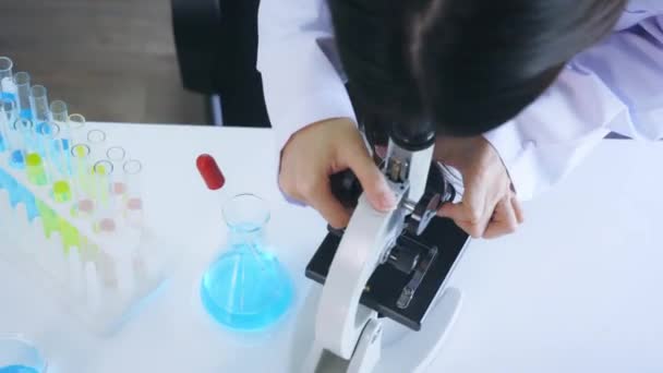 Jovem cientista asiática vestindo labcoat médico realizando pesquisa em laboratório enquanto vê elementos através de microscópio com equipamentos como tubo de ensaio e frasco — Vídeo de Stock