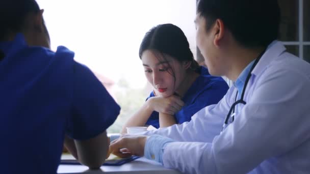Fiatal ázsiai férfi és női orvosok és ápolónők csoportja, akik a szünetben a menzán ülnek és a súlyos kórházi betegeseteket vitatják meg — Stock videók