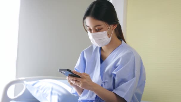 Молодая азиатская пациентка сидит на больничной койке в хирургической маске для безопасности, используя смартфон для смс и просмотра интернета — стоковое видео