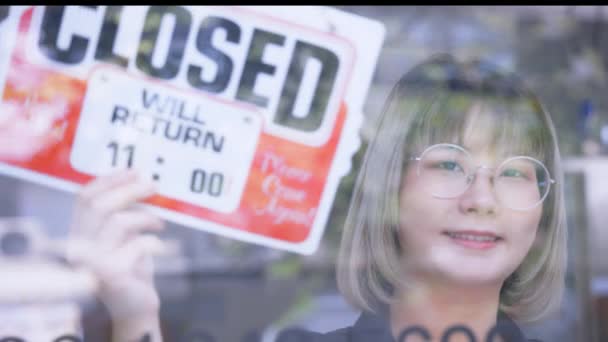 Jeune femme asiatique retournant de signe fermé à signe ouvert par fenêtre en verre. Femme propriétaire de magasin tenant un avis ouvert devant le magasin — Video