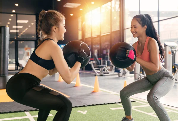 Happy young female athletic people performing squat exercises with friend and holding a medicine ball at fitness gym. Group of two confident women with healthy lifestyle working out together.