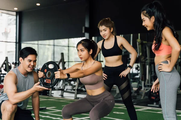 Grupp av människor hejar på sin asiatiska kvinnliga vän gör knäböj med en viktskiva i gym. Arbeta tillsammans som ett lagarbete. — Stockfoto