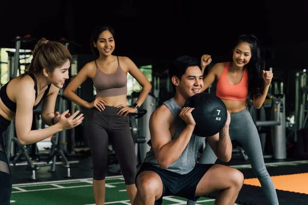 Een groep mensen juichen hun Aziatische Chinese mannelijke vriend toe die hurkt met een medicijnbal in de fitnessruimte. Samenwerken als een teamwork. — Stockfoto