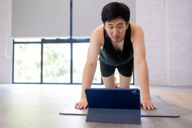 Genç adam tabletten video dersi izlerken evde egzersiz yapıyor. Erkek fitness elemanı içeride çalışıyor.