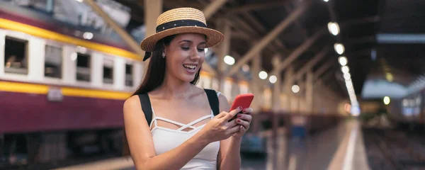 Young brunette woman traveler laughs while playing a mobile phone in train station platform. — Stock Photo, Image