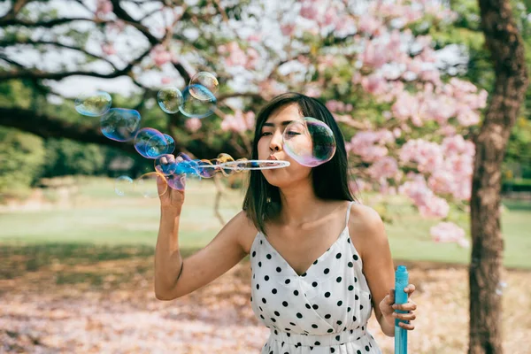 Jovem do Sudeste Asiático étnico 20 anos mulher soprando bolhas de ar no parque. Menina feliz usa vestido de verão com árvores no fundo. — Fotografia de Stock