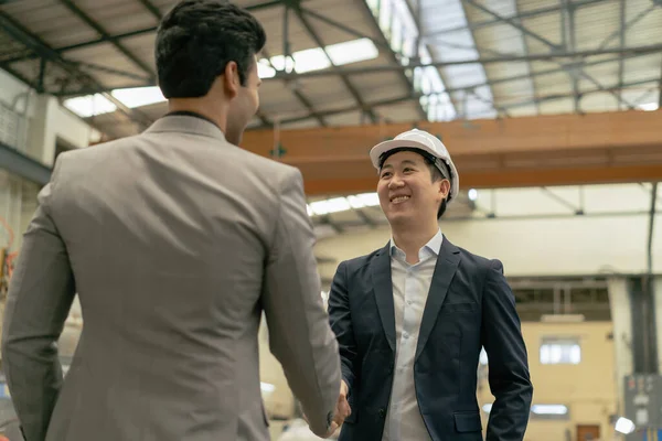 30s young Asian businessman in formal suit and hard hat showing foreign business partner in factory background. Business partnership and team work success concept