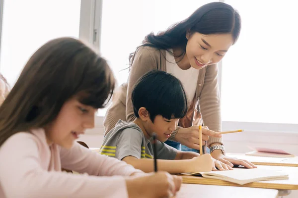 亚洲学校的老师在课堂上帮助学生.在学校工作的年轻妇女帮助男孩写作、教育、支持、照料 — 图库照片