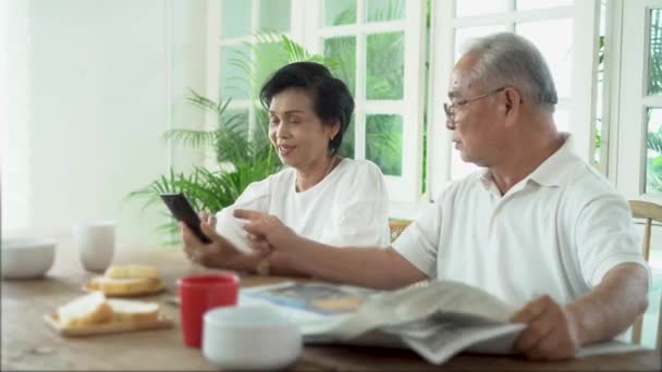 Pareja asiática mayor desayunando juntos en el comedor. 70 jubilado anciano hombre leyendo periódico mientras que la mujer usando el teléfono móvil. — Vídeo de stock