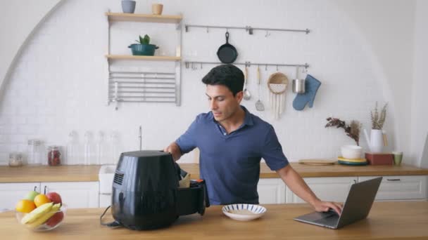 Hombre usando freidora y portátil en la cocina — Vídeos de Stock