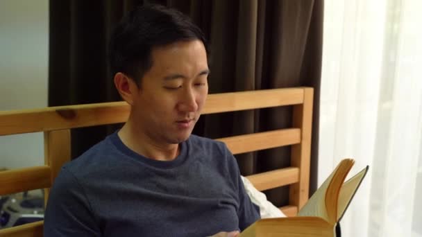 Porträtt av fridfulla 30-talets asiatiska man i vardagskläder tillbringar fritiden med att läsa en bok hemma på sängen. Rekreation och fritid — Stockvideo