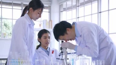 Laboratuvarda birlikte çalışan bir grup genç Asyalı bilim adamı mikroskop kullanarak laboratuvar önlüğü giyiyor.