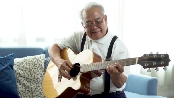 Asiatico anziano uomo seduto sul divano e suonare la chitarra acustica a casa — Video Stock