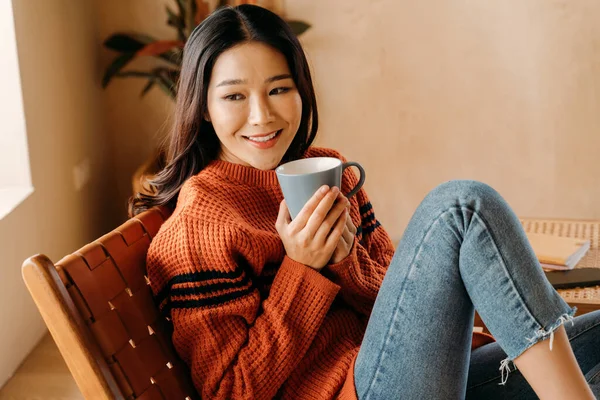 若い美しいアジアの女性は、コーヒーとマシュマロのカップを上に持ち、家に座って窓の外を見ています。寒い冬にセーターでチョコレートを飲む幸せな女の子. — ストック写真