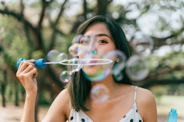 Jovem do Sudeste Asiático étnico 20 anos mulher soprando bolhas de ar no parque. Menina feliz usa vestido de verão com árvores no fundo. — Fotografia de Stock