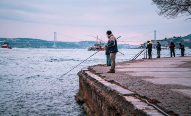Bebek, İstanbul, Türkiye - 19 Şubat 2021 - İstanbul 'un Bebek kentindeki Boğaz' da balıkçılık yapan balıkçılar ve arka planda 15 Temmuz Şehitler Köprüsü