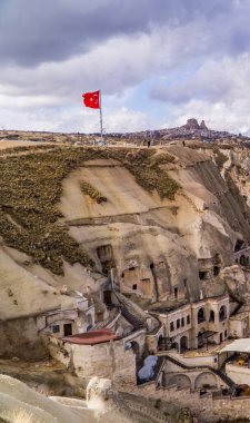 Goreme, Türkiye - 21 Mart 2021 - Türkiye 'nin Kapadokya kentindeki Goreme kasabasının dikey görüntüsü, bacaları, evleri ve Türk bayrağıyla benzersiz kaya oluşumları