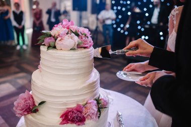 Gelin ve damat ellerinde bıçakla şık beyaz düğün pastasını çiçeklerle kesiyorlar. Pembe ve beyaz şakayıklı modern büyük düğün pastası. Restoranda lüks yemek servisi. düğün resepsiyonu
