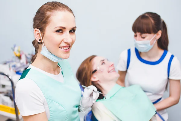 Joven ortodoncista hermosa sonriendo con máscara en el cuello y los instrumentos dentales en la mano con asisstant y paciente en el fondo después de la operación —  Fotos de Stock