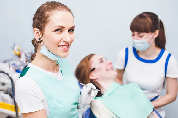 Young beautiful orthodontist smiling with mask on her neck and dental instruments in hand with asisstant and patient on background after operation Telifsiz Stok Fotoğraflar