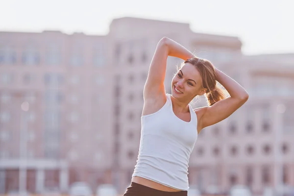 Jeune femme sportive souriante en chemise de sport regardant loin tout en étirant les mains sur le terrain en face de son campus universitaire — Photo