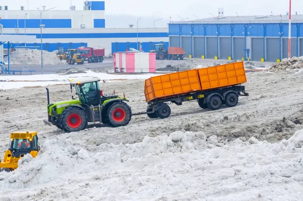 A powerful tractor with a trailer transports snow from city streets to snow-melting clusters of the installation