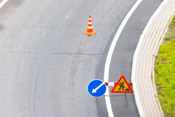 Turn left sign on the road. Warning signs and orange safety traffic cones on background. Direction of detour, roadworks. Road signs denoting road repairs