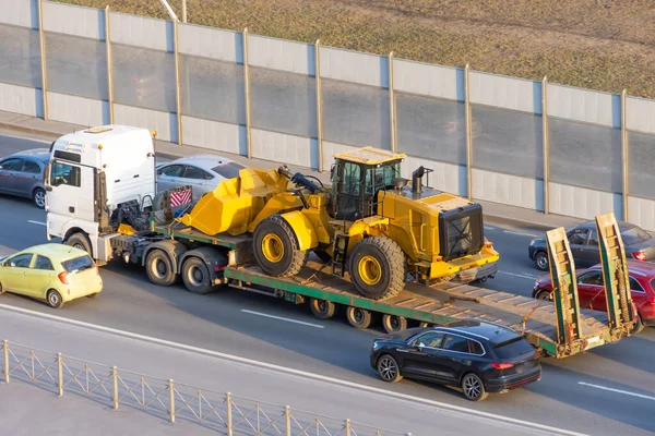 Truck with a long trailer platform for transporting heavy machinery, loaded tractor with a bucket. Highway transportation traffic jam