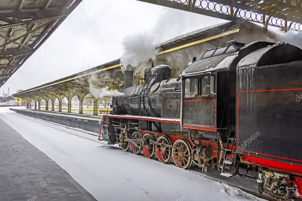 Steam locomotive stands on the platform of the station, winter cold snowy day