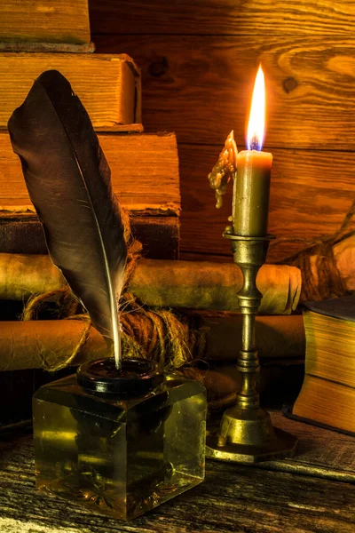 The inkwell with a feather, old books and documents on scrolls, a burning candle stands on an old wooden the table.