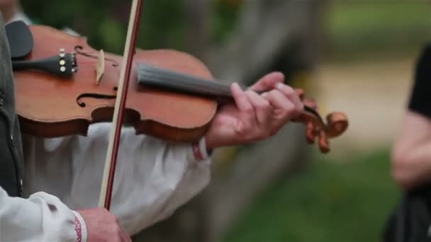 Man in a white traditional shirt playing violin outdoors in street with unfocused musicians and audience on the background. Close up — Αρχείο Βίντεο