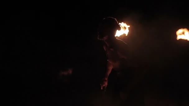 Fire show performance. Handsome male fire performer twirling fire baton and making fire breathing spitting flame against black background. Slow motion — 비디오