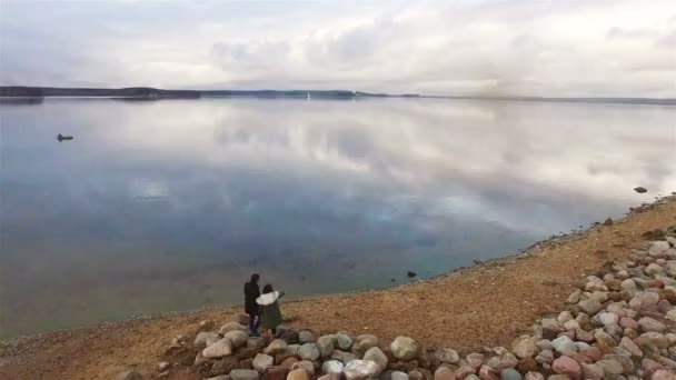4k vuelo aéreo sobre la orilla del lago, donde dos personas amigos o pareja de pie viendo magníficas nubes sobre la bahía del lago tranquilo o el río y señalando la planificación de algo — Vídeo de stock