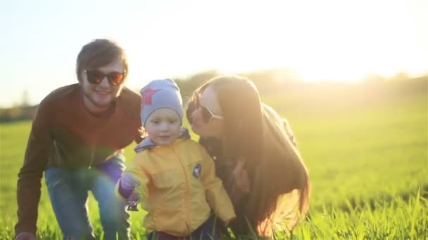 Jovens pais caucasianos saudáveis fazendo surpresa e abraçando o menino da parte de trás rindo em um prado ao pôr do sol da noite com sol retroiluminado e grama verde da primavera no fundo — Vídeo de Stock