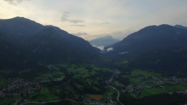 Mountain valley town with houses near river 4k aerial birds eye view from above. Fly over small town in Switzerland at river valley in morning or evening sunset or sunrise time clouds weather — ストック動画