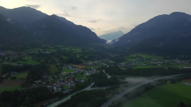 Mountain valley town with houses and road near river 4k aerial bird eye top view from above. Fly over small town in Switzerland and local road with traffic cars river valley morning evening fog clouds — Stock Video