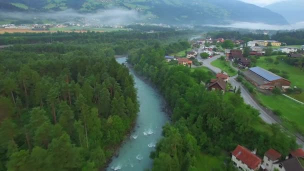 Dağ Nehri Vadisi şehir evleri ve yol 4k hava kuş bakışı yukarıda. İsviçre sabah akşam zaman sis bulutları çatı üstüne Nehri Köprüsü köy binalar ile güneş enerjisi panelleri üzerinde uçmak — Stok video