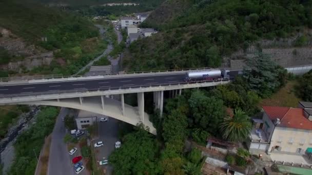 Mountain road overpass bridge up sea resort coastal town 4k panoramic aerial drone view from above. Fly over transport traffic with cars and tank trucks road railroad junction Italy or France Europe — ストック動画