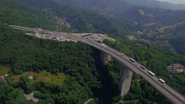Overpass viaduct bridge road with truck lorry cars move aerial 4K. Fly over mountain bypass highway with transport traffic and parking lot above green forest valley town. Logistics in Italian Alps — ストック動画