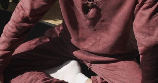 Seguimiento de disparo joven yogui sentado en asana de loto meditando en el interior de cámara lenta. Primer retrato detallado del hombre barbudo sentado con las piernas cruzadas con las manos de Namaste fondo soleado. Yoga relajación zen — Vídeos de Stock