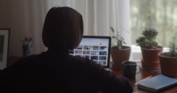 Rückenansicht eines Mädchens, das am Tisch sitzt und zu Hause mit einem Notizbuch in Zeitlupe arbeitet. Nicht wiederzuerkennende Freiberuflerin sucht Shutterstock-Bilder auf Laptop-natürlichem Licht. Selbstständiges Mikrostock-Geschäft — Stockvideo