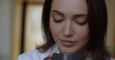 Kapalı mekanda kahve içen zarif bir kadının portresi. Doğal hafif, ağır çekim. İyi giyimli bir kız elinde çay fincanıyla sığ arka planda kameraya bakıyor. Sabah kahvaltısı konsepti