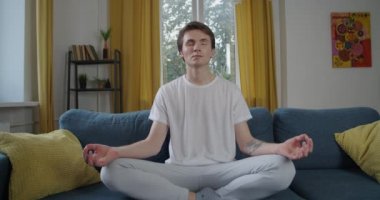 Genç bir adamın evde tek başına meditasyon yapması doğal ışık yavaş çekim. Çekici, gözleri kapalı bir insan kanepeye oturur ve yoğun bir haftanın arka planında dinlenir. Stres karşıtı yaşam tarzı